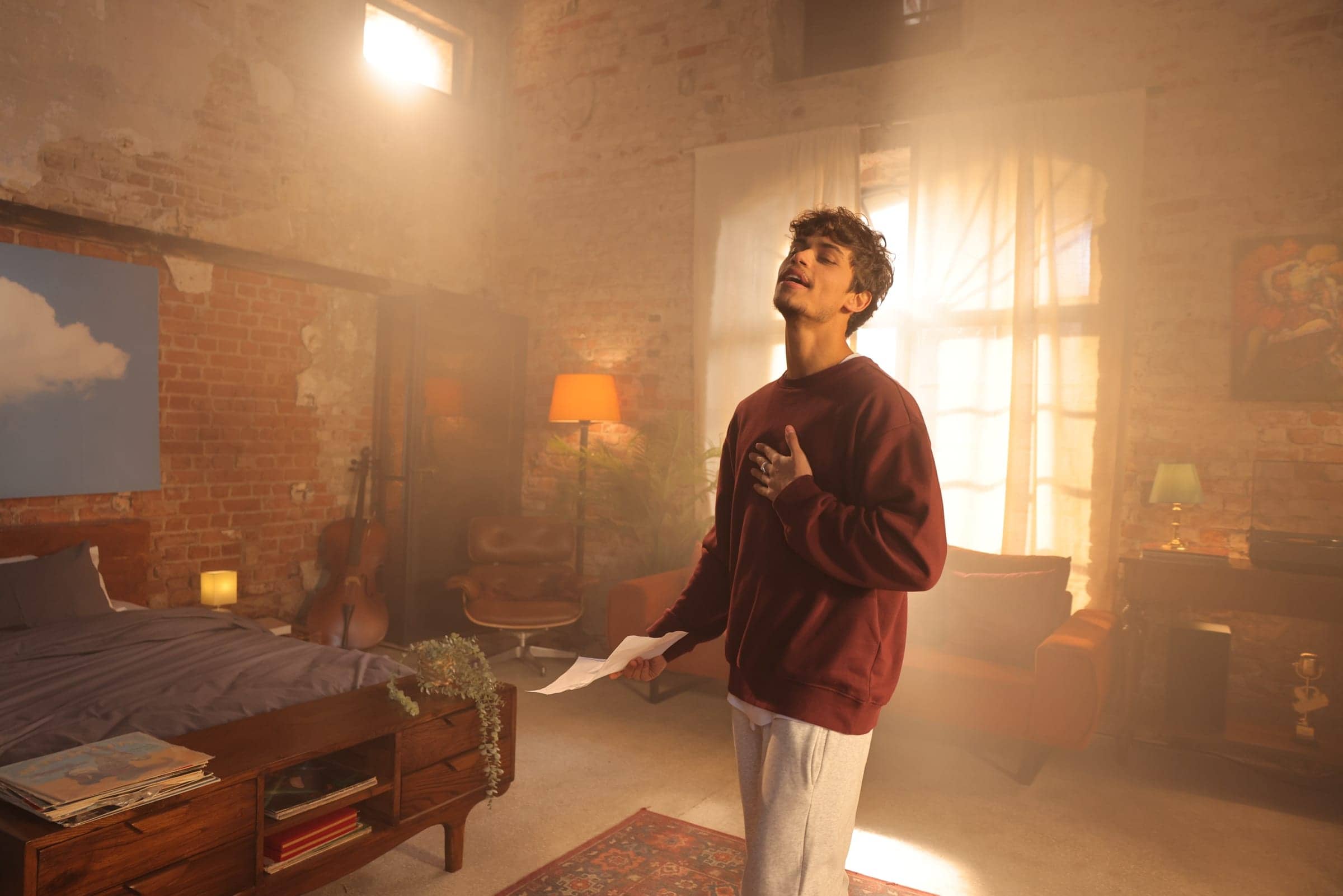 Harris J singing with his hand on his heart, eyes closed, in a sunlit room