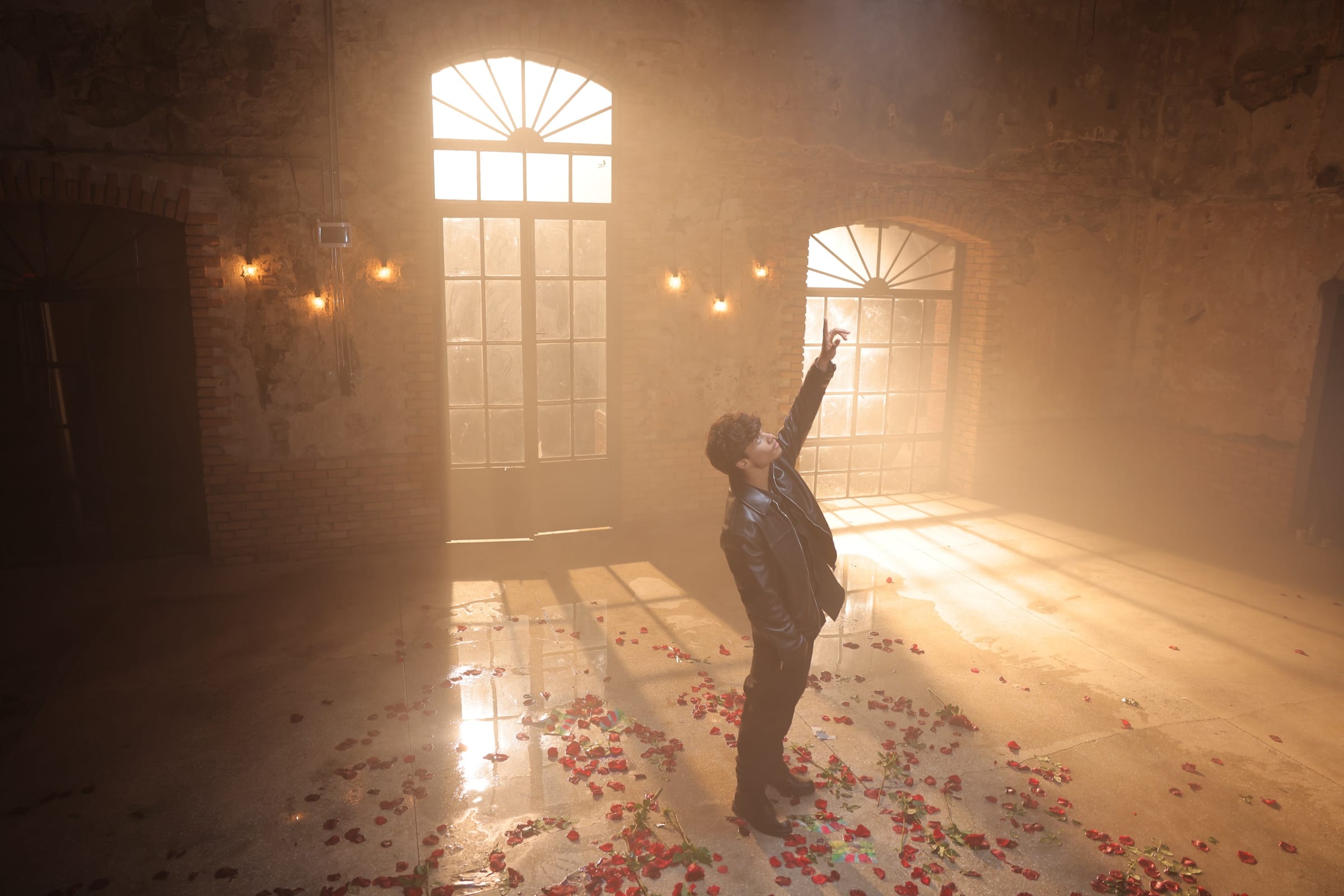 Harris J reaching upwards in a sunlit brick hall, rose petals scattered across the floor